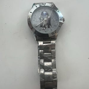 May include: Silver-toned wristwatch featuring a Star Wars R2-D2 graphic on the watch face. The watch has a stainless steel band and a round face with Roman numeral hour markers. The watch case is approximately 1.5 inches in diameter.