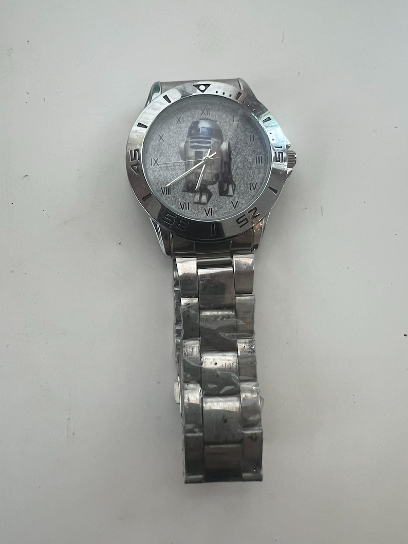 May include: Silver-toned wristwatch featuring a Star Wars R2-D2 graphic on the watch face. The watch has a stainless steel band and a round face with Roman numeral hour markers. The watch case is approximately 1.5 inches in diameter.