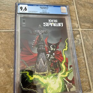 May include: A graded comic book titled "King Spawn #1" with a 9.6 grade. The cover art features a dark figure with a red cape, a large cross-shaped weapon, and the title "King Spawn". The comic is encased in a clear protective holder.