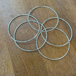 May include: Five silver bangles with a textured, sparkling design are arranged on a wooden surface. The bangles are circular and appear to be made of metal. The light reflects off the textured surface of the bangles.
