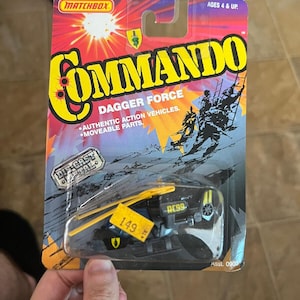 Puede incluir: Un vehículo de juguete Matchbox Commando Dagger Force de metal fundido a presión en su embalaje original. El embalaje es rojo, amarillo y azul con el texto "COMMANDO" y "DAGGER FORCE". El juguete es negro y amarillo.