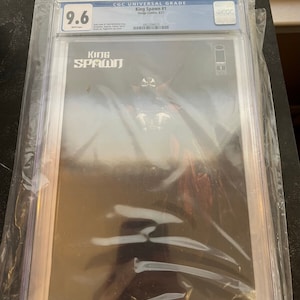 Puede incluir: Un cómic clasificado en una caja de plástico. El cómic es King Spawn #1, clasificado 9.6 por CGC. La portada presenta un personaje negro y rojo con una cara de calavera.