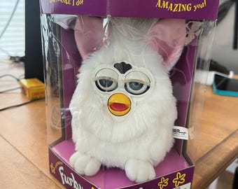 May include: A white Furby toy in its original packaging. The Furby has pink ears, a yellow beak, and blue eyes. The packaging is purple and features the Furby logo and the text "I will KEEP AMAZING you!"