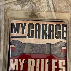 Könnte beinhalten: Eine dekorative Lichtschalterabdeckung mit dem Text "MY GARAGE MY RULES". Die Abdeckung hat einen roten und beigefarbenen Hintergrund mit einer Schraubenschlüsselgrafik. Der Text ist fett und ohne Serifen.
