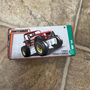 Puede incluir: Una caja de coche de juguete Matchbox con un vehículo todoterreno 4x4 rojo con neumáticos negros y llantas amarillas. La caja es verde y naranja con el logotipo de Matchbox y el texto "MBX 4x4".