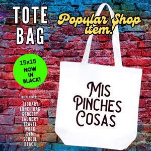 May include: White tote bag with black text that reads "Mis Pinches Cosas". The bag is on a brick wall background with a green circle that says "15x15 Now In Black!" and a list of uses for the bag.