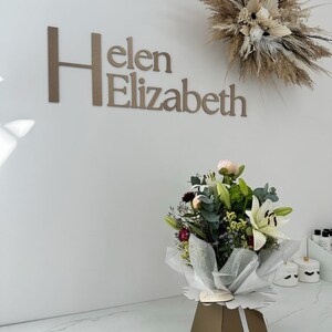 May include: A wall sign with the text Helen Elizabeth in large brown letters. Above the sign is a dried floral arrangement. Below, a bouquet of white lilies, pink peonies, and greenery is wrapped in white translucent paper in a brown box.