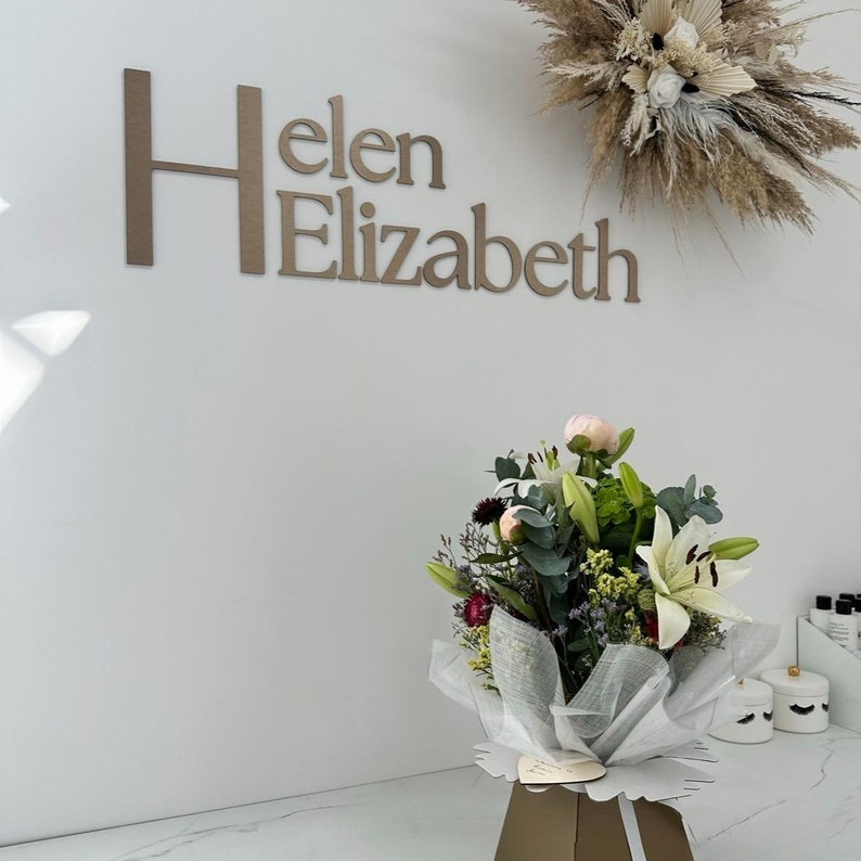 May include: A wall sign with the text Helen Elizabeth in large brown letters. Above the sign is a dried floral arrangement. Below, a bouquet of white lilies, pink peonies, and greenery is wrapped in white translucent paper in a brown box.