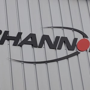 May include: A three-dimensional sign featuring the word SHANNON in bold black capital letters mounted on a light gray vertical ribbed wall. The letter O is replaced by a solid red circle, and a thin black curved line sweeps across the text.