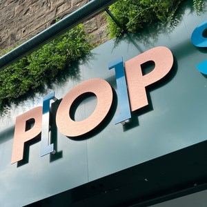 May include: A dark green sign featuring three-dimensional lettering that spells PLOP S. The letters P, O, and P are finished in a metallic copper tone, while the letters L and S are rendered in a glossy blue finish. Green foliage sits above the sign.