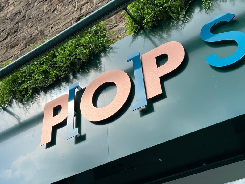May include: A dark green sign featuring three-dimensional lettering that spells PLOP S. The letters P, O, and P are finished in a metallic copper tone, while the letters L and S are rendered in a glossy blue finish. Green foliage sits above the sign.