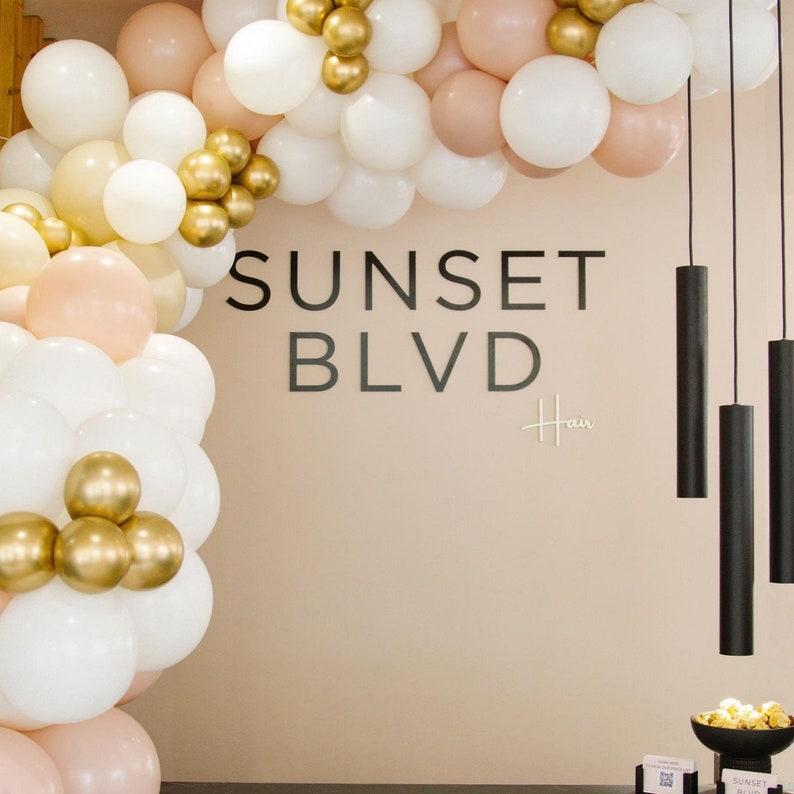 May include: A balloon arch featuring white, peach, and metallic gold balloons framing a wall with the black text SUNSET BLVD Hair. Three black cylindrical pendant lights hang on the right side, and a bowl of gold balloons sits on a stand.
