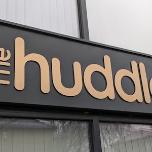 May include: A black rectangular storefront sign featuring the text The huddle in large, three-dimensional, light tan sans-serif letters. The sign is mounted above a glass window pane, with the letters positioned horizontally across the dark surface.