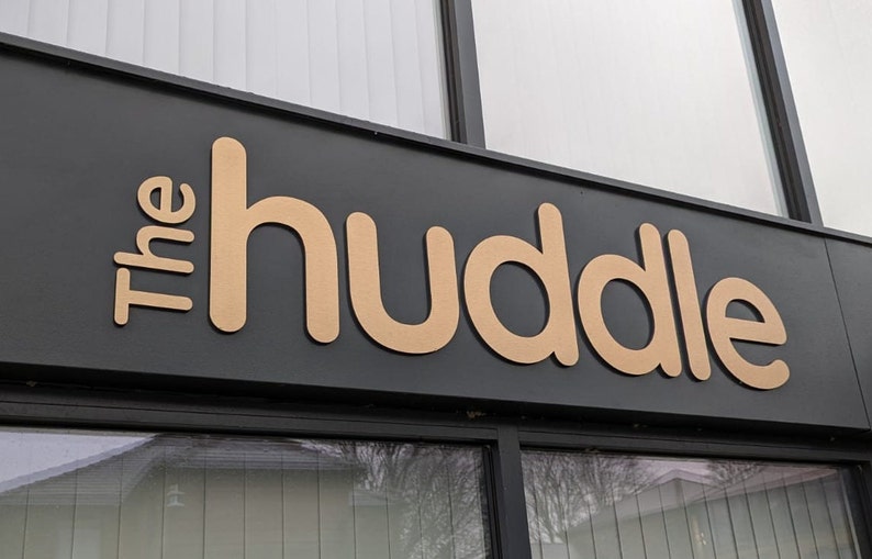 May include: A black rectangular storefront sign featuring the text The huddle in large, three-dimensional, light tan sans-serif letters. The sign is mounted above a glass window pane, with the letters positioned horizontally across the dark surface.