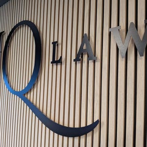 May include: A wall-mounted sign featuring large, black, three-dimensional letters spelling JQ LAW. The letters are mounted on a background of vertical, light-colored wooden slats with narrow, dark gaps between each piece of wood.