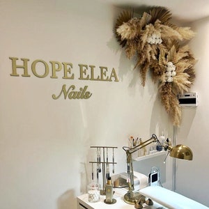 May include: A nail salon station featuring gold wall lettering that reads HOPE ELEA Nails, accented by a large dried pampas grass and white flower arrangement. The setup includes a gold desk lamp, a tool stand, and a white hand rest on a desk.