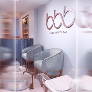 May include: A beauty salon interior featuring two light blue velvet armchairs with vertical channel tufting, copper-toned side tables, and a wall sign displaying the text bbb BELLES BEAUTY BAR in large copper-colored lettering against a pale wall.