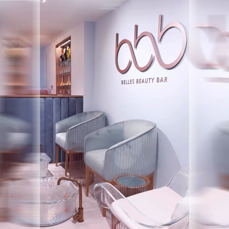 May include: A beauty salon interior featuring two light blue velvet armchairs with vertical channel tufting, copper-toned side tables, and a wall sign displaying the text bbb BELLES BEAUTY BAR in large copper-colored lettering against a pale wall.