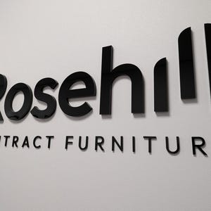 May include: A wall-mounted sign featuring the word Rosehill in large, glossy black, three-dimensional lettering, positioned above the words CONTRACT FURNITURE in smaller, matching black, flat, sans-serif capital letters on a white wall.