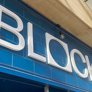 May include: A white sign with the word "BLOCK" in large, bold letters on a blue tiled wall. The sign is illuminated by two light bulbs.