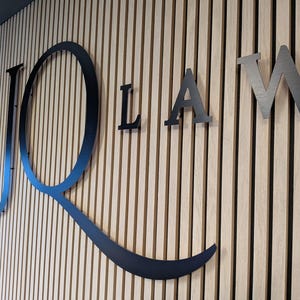 May include: A large, black, metal sign featuring the letters JQ LAW mounted on a wall with vertical light-colored wooden slats. The letter Q is oversized and stylized with a long, sweeping tail that curves underneath the letters LAW.