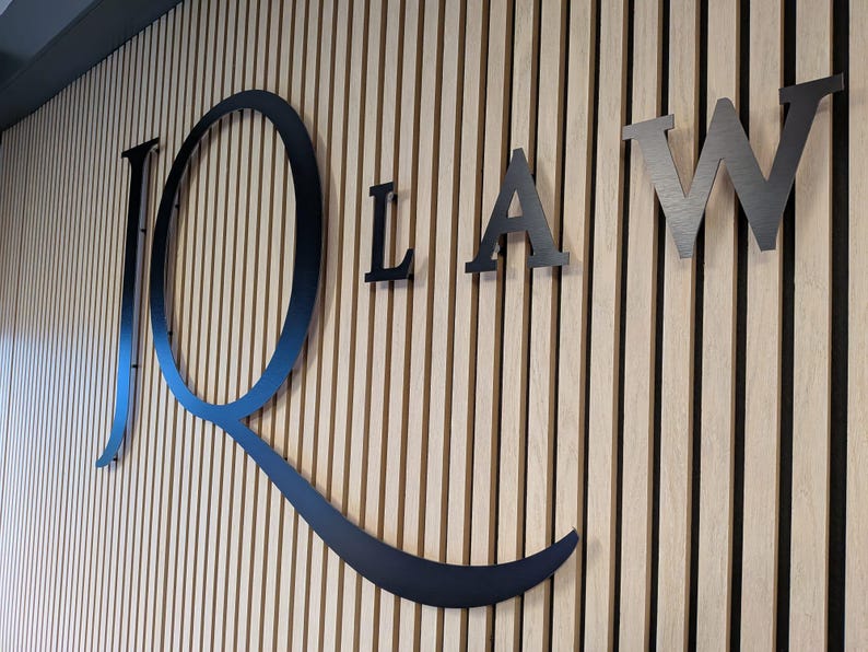 May include: A large, black, metal sign featuring the letters JQ LAW mounted on a wall with vertical light-colored wooden slats. The letter Q is oversized and stylized with a long, sweeping tail that curves underneath the letters LAW.