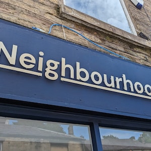May include: A dark blue storefront sign featuring the word Neighbourhood in large, cream-colored, three-dimensional sans-serif letters. The sign is mounted on a dark blue wooden panel above a window, with a thin cream line underlining the text.