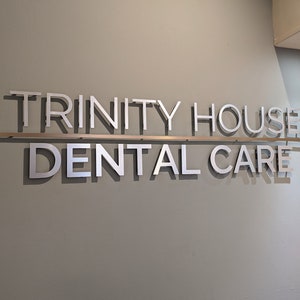 May include: A wall-mounted sign featuring the silver-colored text TRINITY HOUSE DENTAL CARE in a sans-serif font. The words are arranged on two lines, separated by a thin horizontal metal bar, casting distinct shadows against the light gray wall.