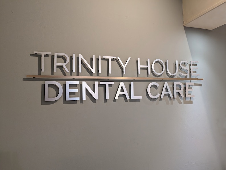 May include: A wall-mounted sign featuring the silver-colored text TRINITY HOUSE DENTAL CARE in a sans-serif font. The words are arranged on two lines, separated by a thin horizontal metal bar, casting distinct shadows against the light gray wall.