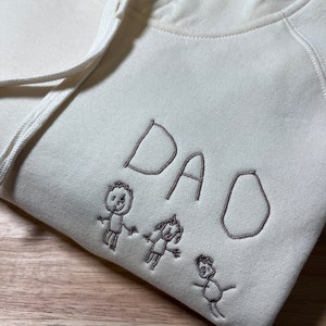 Pode incluir: Um moletom branco com um design bordado marrom da palavra "DAD" e tr&ecirc;s figuras de palitos de m&atilde;o dadas.