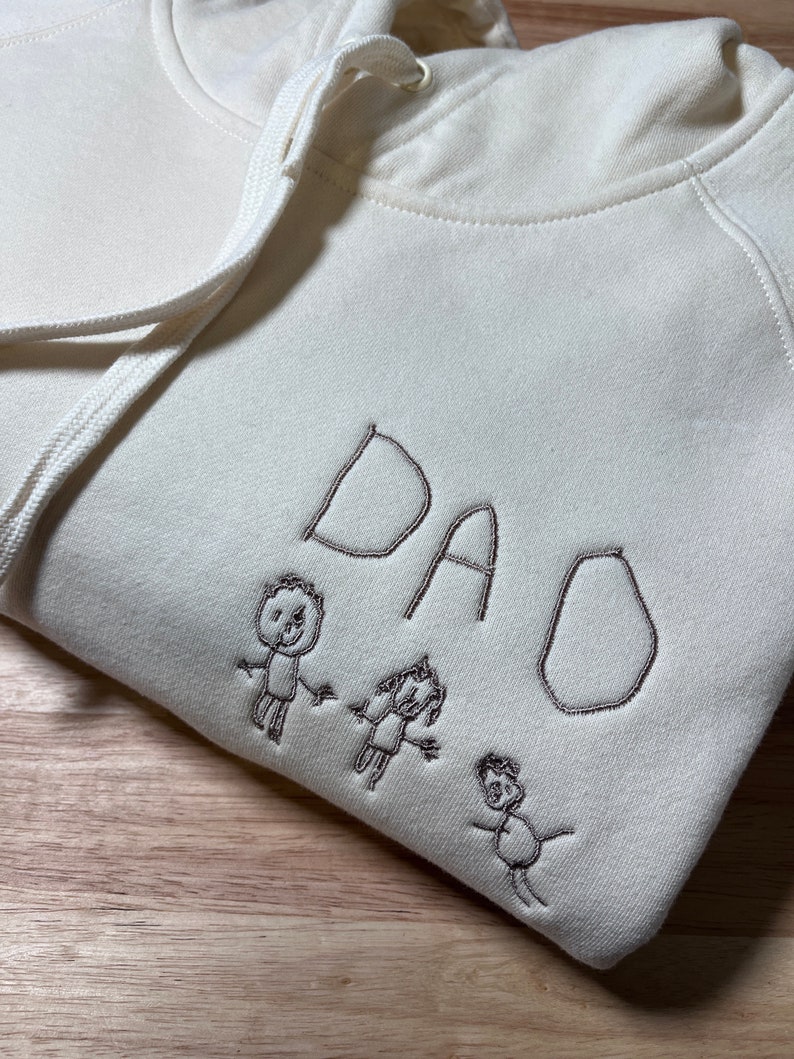 Pode incluir: Um moletom branco com um design bordado marrom da palavra "DAD" e tr&ecirc;s figuras de palitos de m&atilde;o dadas.