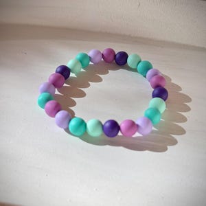 Könnte beinhalten: Ein farbenfrohes Perlenarmband mit einer Mischung aus runden Perlen in Lila-, Türkis- und Lavendeltönen. Das Armband besteht aus glatten, massiven Perlen und ist zum Tragen am Handgelenk konzipiert. Das Armband ist ein Modeaccessoire.