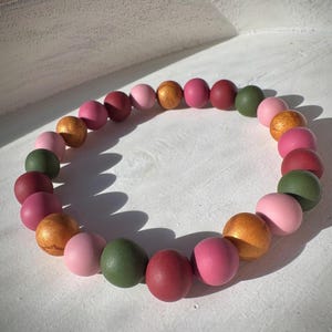 May include: A beaded bracelet composed of various colored wooden beads. The beads are in shades of pink, red, green, and gold. The bracelet is circular and rests on a white surface.