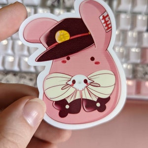 May include: A pink bunny rabbit wearing a black and gold officer's hat with a red and white band. The bunny is wearing a white bow tie with black accents and has a skull and crossbones design on its chest.