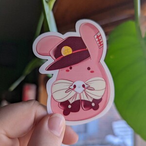 May include: A pink bunny rabbit wearing a burgundy hat with a gold button and a white bow tie. The bunny has a red nose and black eyes.