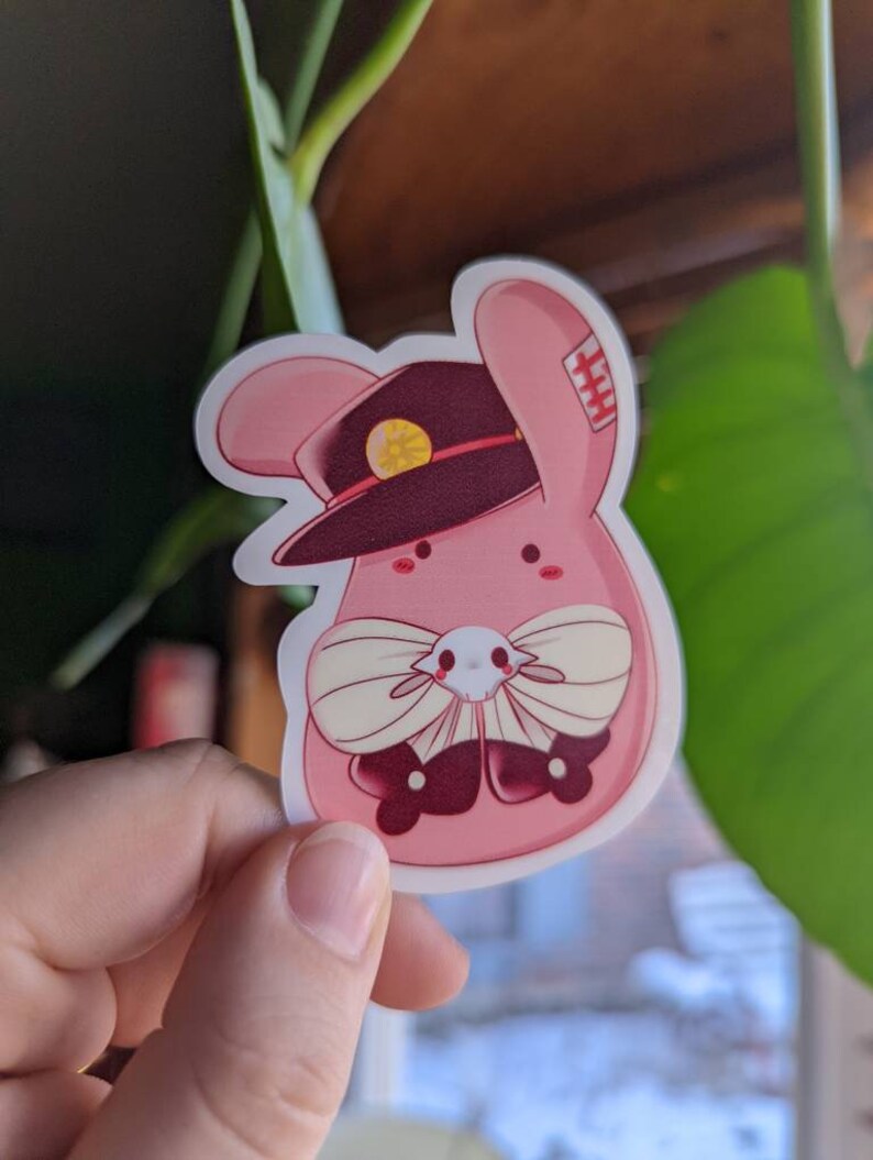May include: A pink bunny rabbit wearing a burgundy hat with a gold button and a white bow tie. The bunny has a red nose and black eyes.
