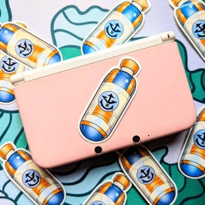 May include: A pink Nintendo 3DS handheld gaming console with a blue and orange sticker of a bottle with a white anchor symbol on it. The sticker is on the top screen of the console.