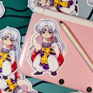 May include: A sticker featuring a cartoon character with white hair, a crescent moon on their forehead, and a yellow and purple sash. The character is wearing a white and red outfit with a furry white collar. The sticker is on a pink surface.