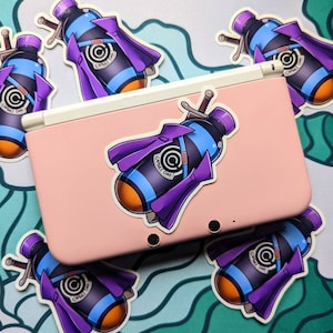 May include: A pink Nintendo 3DS XL console with multiple stickers of a blue and orange capsule with a purple cape and the text "Capsule Corp" on it.