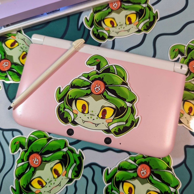 K&ouml;nnte beinhalten: Ein rosa Nintendo 3DS Handheld-Spielkonsole mit einem wei&szlig;en Stift und einem Aufkleber mit einer Cartoon-Medusa mit gr&uuml;nen Haaren und gelben Augen. Mehrere &auml;hnliche Aufkleber sind um die Konsole herum verteilt.