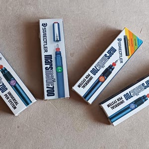 Op de afbeelding: Vier Staedtler Marsmatic 700 technische pennen in hun originele witte kartonnen verpakking. Elke doos toont de merknaam, het model en een afbeelding van de pen met een gekleurde ring die de specifieke lijndikte van de punt aangeeft.