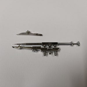 May include: A silver-toned metal drafting compass set consisting of a main tool with an adjustable needle point and a separate interchangeable attachment. The main component features a central rod, multiple adjustment screws, and a sharp point.