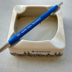 Puede incluir: Un lápiz mecánico azul con el texto "787 MARS-PAN-TECHNICO" impreso en el lateral, descansando en un cenicero de cerámica blanca con un diseño de ciudad en blanco y negro y el texto "STAEDTLER" impreso en la parte inferior.