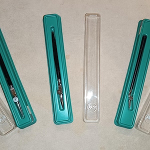 May include: Several black Kern drawing pens in teal cases with clear plastic covers. The pens are in various stages of being opened. The Kern logo is visible on the clear plastic cases. The pens are on a light brown surface.