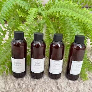 May include: Four brown glass bottles with black caps, each with a white label reading "The Diffuser Flower Co. Luxury fragrance oil". The bottles are displayed against a backdrop of green fern leaves.