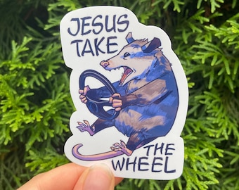 May include: A white sticker with a cartoon possum driving a steering wheel. The sticker says "Jesus Take The Wheel" in black text.