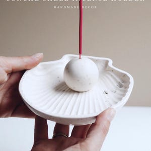 Op de afbeelding: Een witte schelp wierookhouder met een bolvormige houder en een rode wierookstok. De houder is handgemaakt en heeft een textuur. De tekst "CONCHÉ SHELL INCENSE HOLDER HANDMADE DECOR" is zichtbaar.