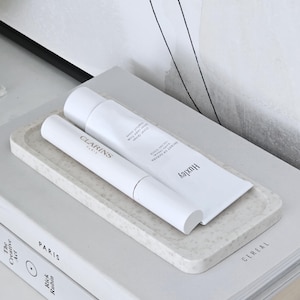 May include: A speckled white Vero Flat Tray with two white cosmetic tubes, one Clarins and one Huxley. The tray sits on a stack of books, including "Paris" and "The Creative Act". The background features an abstract design.