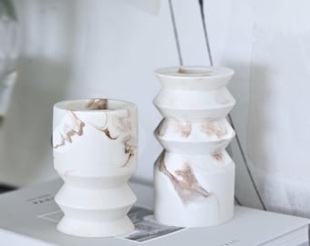 Pode incluir: Dois castiçais de cerâmica branca com marmoreio castanho. Um é um cilindro curto e largo, e o outro é um design mais alto e empilhado. O texto "CRANTE CANDLE HOLDER" e "HANDMADE DECOR" são visíveis. Os castiçais estão em uma pilha de livros.