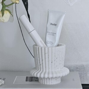 May include: A white speckled ceramic decorative holder with a fluted design, holding a white tube of Huxley hand cream and a white tube of Clarins product. The holder sits on a stack of books with the word "PARIS" visible.